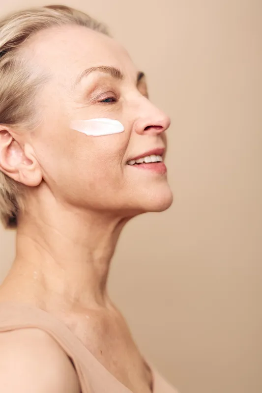 Smiling mature woman with wrinkles applies white face cream on cheek in close-up profile. Concept of skincare, aging beauty, facial care, wellness, and natural self-care routine for healthy skin.