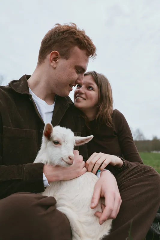 Joyful couple outdoors with a playful goat kid, sharing tender moments in a grassy pasture. Warm natural light, setting convey countryside lifestyle and animal companionship and warmth.