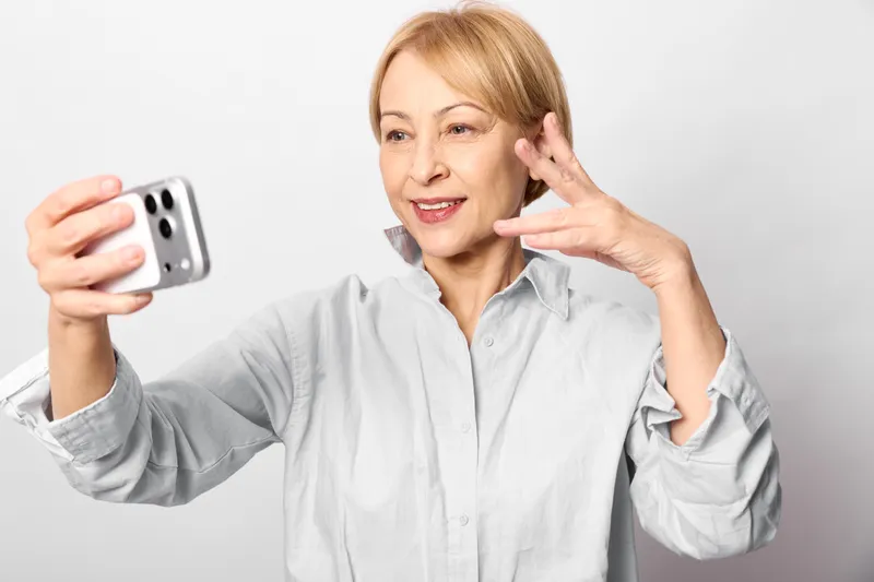 Elderly woman smartphone selfie technology communication social portrait, smiling and touching her ear, holding phone indoors, concept of mobile connection