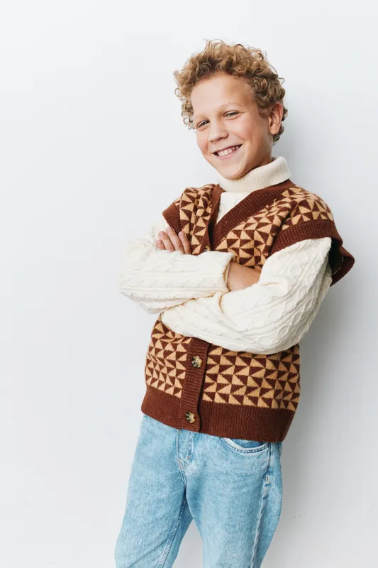 A cheerful young boy with curly hair smiles confidently while leaning against a plain wall, wearing a cozy geometric brown cardigan and a cream knit sweater.