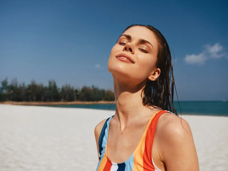 Closeup of a joyful woman on a sunlit beach, eyes closed, basking in warm light as waves meet soft sand under a clear blue sky.