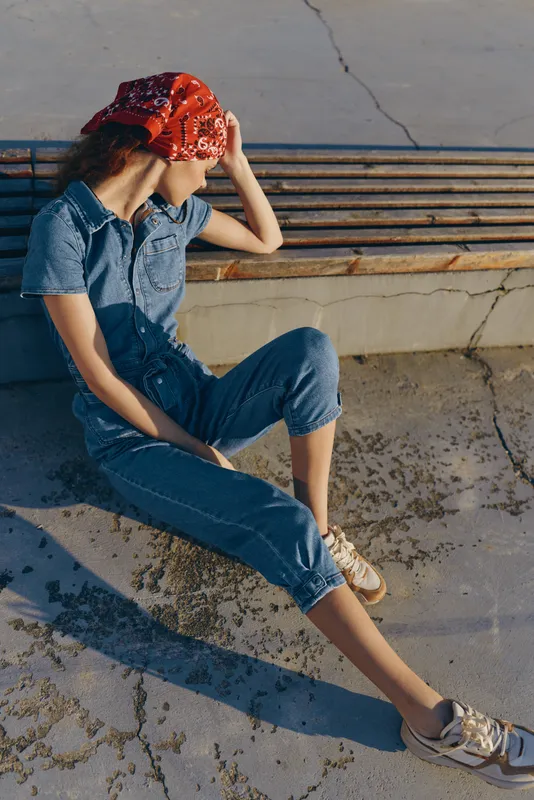 Woman in denim jumpsuit and red bandana sitting on ground by wooden bench, relaxed pose, casual outfit in sunny outdoor urban setting, natural light