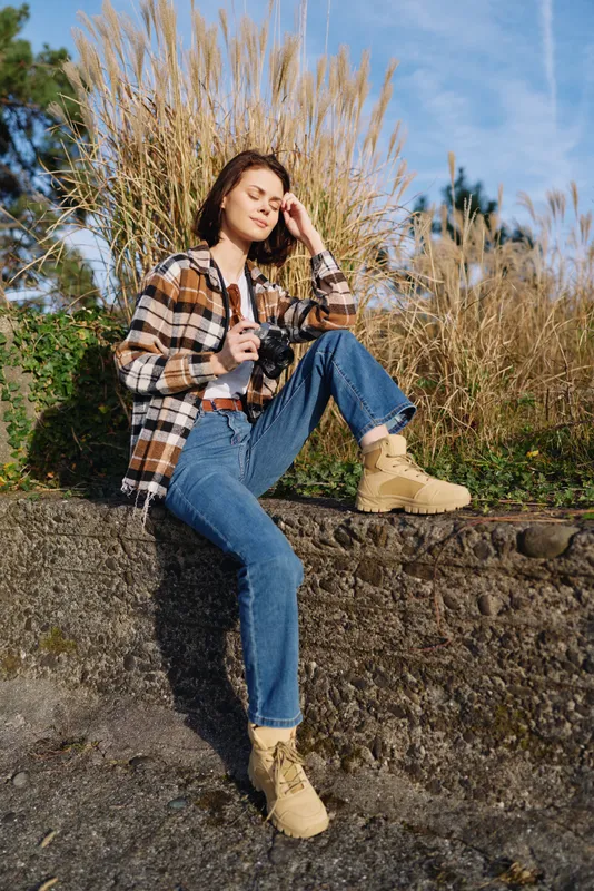 Camera, outdoor, woman, casual, plaid, denim, boots, nature, sunlight, photography portrait lifestyle fashion with a relaxed sitting pose on a stone ledge under a bright sky