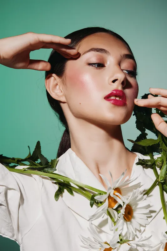 Young woman with glossy red lips and glowing skin poses with hands near face, wearing white and surrounded by chamomile flowers. Studio shot on mint green background. Beauty fashion concept.