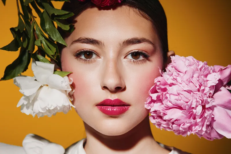 Close-up portrait of a young woman with vibrant pink lipstick holding white and pink peony flowers near her face on a contrasting yellow background. High fashion beauty concept.