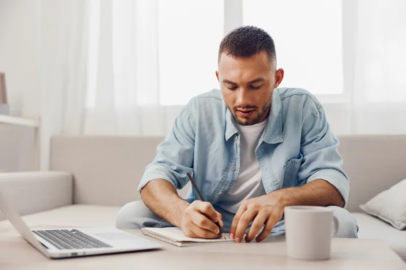 Focused man working at home, writing in a notebook while sitting comfortably in a cozy living room with a laptop. Ideal for modern lifestyle themes in 2025.