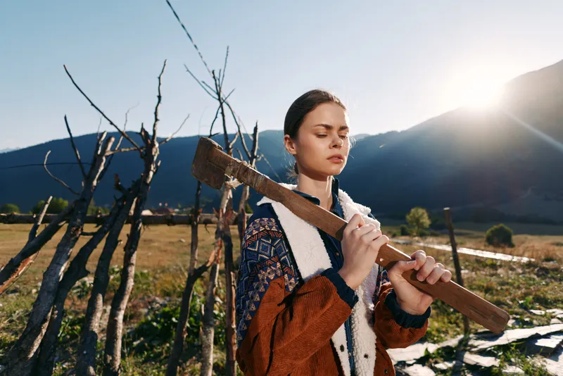 woman with axe in countryside mountains outdoors, portrait of a serene young female holding a vintage axe by a rustic fence with sun flare, autumn jacket and closed eyes