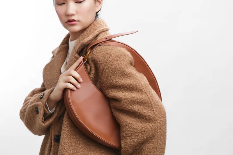 asian woman wearing a chic coat and carrying a leather bag stands in a clean studio, radiating modern style, poise, and quiet confidence for a fashion shoot.