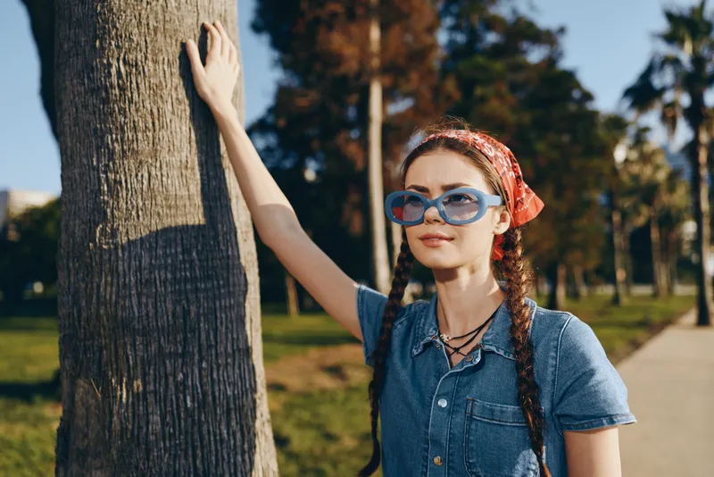 Lifestyle woman in modern boho-western denim street style wearing blue sunglasses and red bandana by tree, casual outdoor summer look with film color effect, young and stylish.