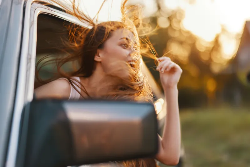 Young woman enjoying a breezy summer drive, wind in her hair, carefree and joyful expression, natural light, sunny background, adventure theme.