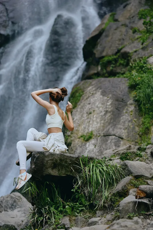 woman meditating near waterfall in white activewear, sitting on rock surrounded by lush greenery, peaceful outdoor nature scene promoting wellness and relaxation