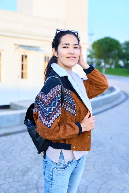 Woman wearing a brown patterned bomber jacket and jeans with a backpack, standing on a sunny street, adjusting her hair, casual urban fashion in a relaxed outdoor mood with soft light.