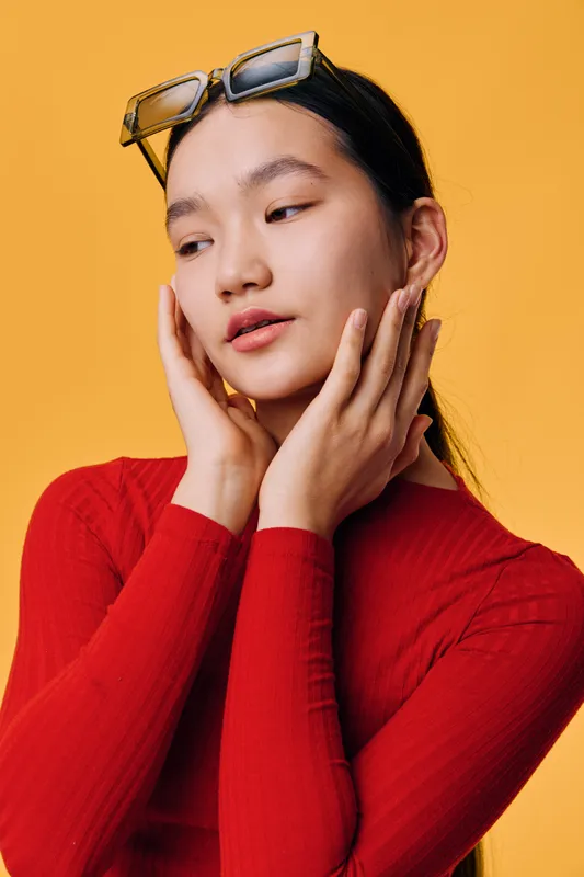Young woman with glasses on head wearing red sweater posing with closed eyes and hands near face on orange background studio portrait of female model with smooth skin and natural look
