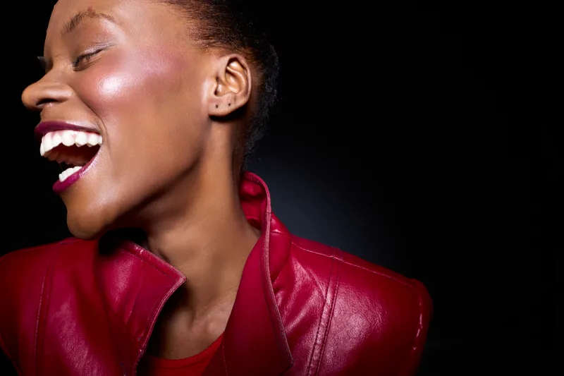 Close-up of a joyful African woman with dark skin laughing, wearing a red leather jacket against a black background. Concept of happiness, fashion, and vibrant energy.