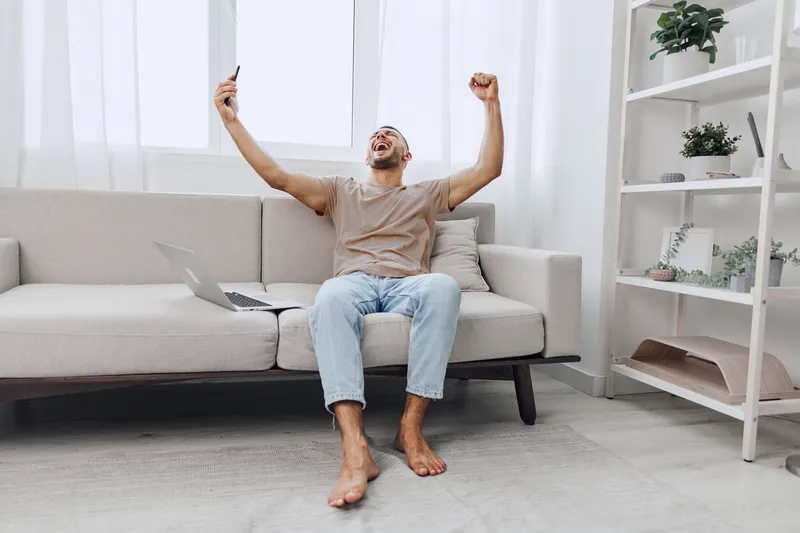 Excited man celebrating a victory, expressing emotions through his joyful gestures while sitting at home, embracing digital experiences and personal milestones. ###