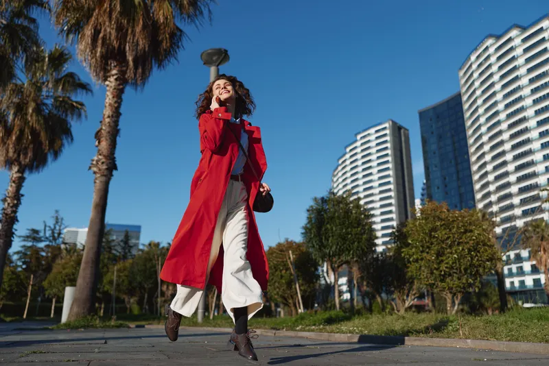 Young woman in a red coat strides along a sunny city street with modern buildings behind, palm trees framing the scene and a confident, playful expression.
