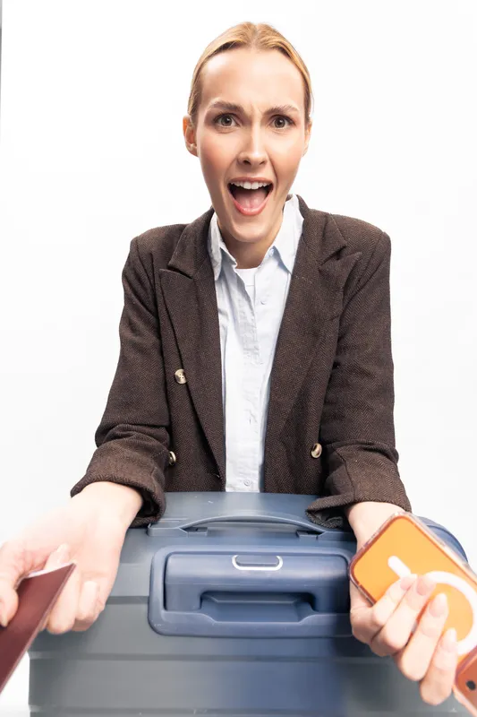 Excited woman in brown jacket holds passport and orange luggage tag standing behind blue suitcase. Travel concept with happy female traveler preparing for trip isolated on white background.