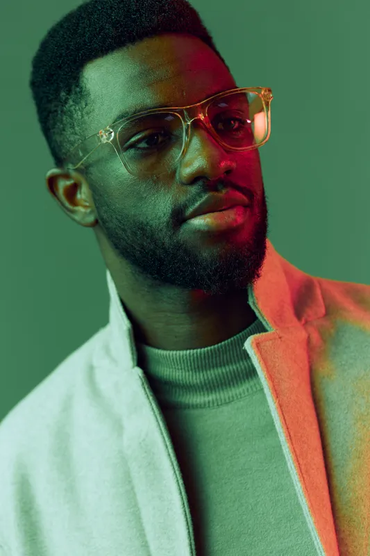 Stylish young Black man in glasses wearing a neutral outfit against a vibrant green background, exuding confidence and modernity