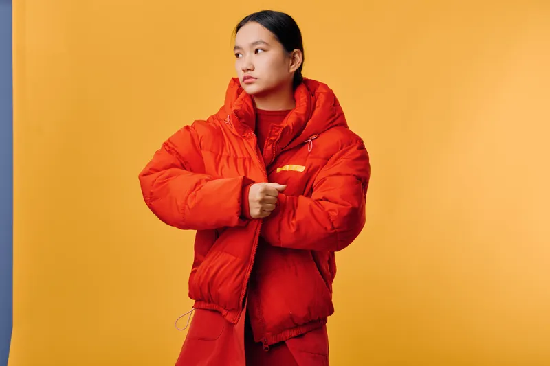 Woman wearing bright red puffer jacket and matching outfit posing against yellow background. Stylish Asian female model in winter clothes looking sideways in fashion photography studio scene.