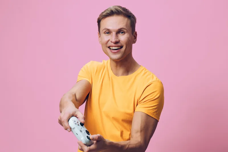 Cheerful man with short hair wearing a bright yellow t-shirt holding game controller, smiling and having fun indoors against a pink background. Happy lifestyle concept, recreation, entertainment, male