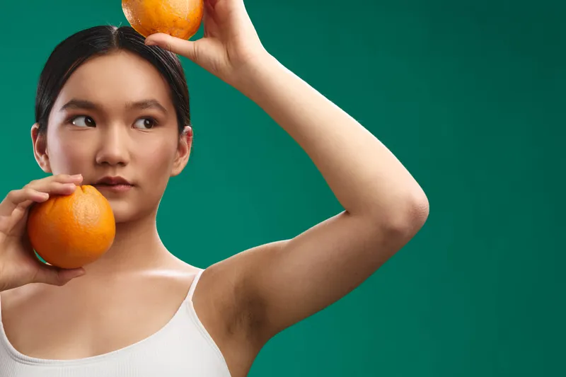 asian woman posing with oranges in a vibrant studio setting; she faces slightly to the side, balancing fruit above her head, creating a fresh and playful mood with bold color contrast.