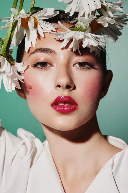 Close-up of young woman with fresh blush and glossy red lips posing with white flower petals on face. Soft LED studio lighting emphasizes beauty and fashion concept.