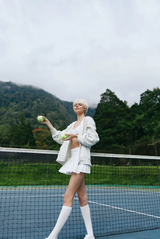 Young female tennis player in white sportswear, smiling with tennis balls on a cloudy day, surrounded by lush green landscape, perfect for sports themed visuals