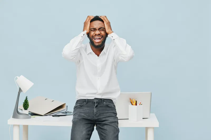Confused man in white shirt holding head, standing behind modern office desk with open notebook, potted plant, and stationery, expressing frustration or stress, isolated on light blue background. Peop