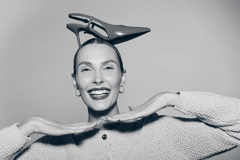 Happy white woman with light skin wears high heel shoe on head and smiles, posing with hands under chin. Concept of humor, fashion, and playful mood.