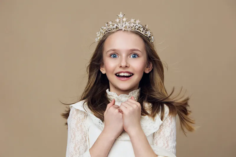 A cheerful young girl in a delicate white dress and tiara, radiating joy against a neutral beige background, perfect for themes of childhood happiness