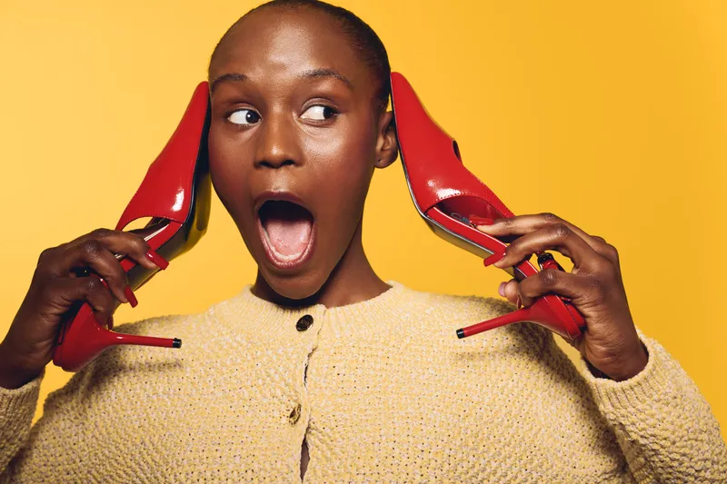 Surprised black woman with dark skin holds red high heels near ears, wearing beige sweater. Bright yellow background emphasizes expressive face and playful concept of fashion and fun.
