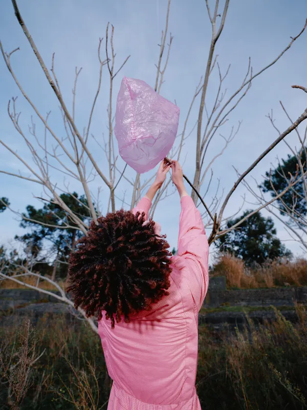 Outdoor pink scarf woman reaching toward a leafless tree under a bright sky, capturing nature, motion and calm in a vivid field scene with soft light and wind
