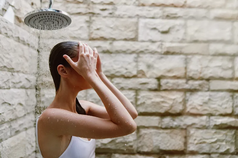 woman showering outdoors under rain showerhead, wet hair, natural stone wall background, calm and refreshing moment in natural light bathing scene