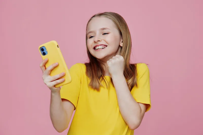 Cheerful young girl celebrating success while using a smartphone, wearing a yellow shirt against a pink background, expressing joy and excitement, perfect for social media themes