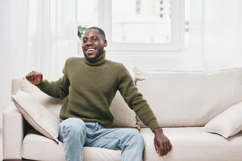 Smiling African man in a cozy green sweater sitting on a light sofa, embodying joy and comfort in a bright living room with natural light