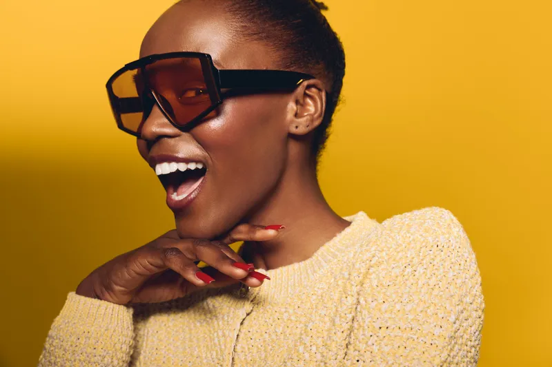Happy African woman with dark skin wearing large sunglasses and yellow sweater laughs joyfully. Studio portrait with bright yellow background. Concept of fashion, style, and positive emotions.