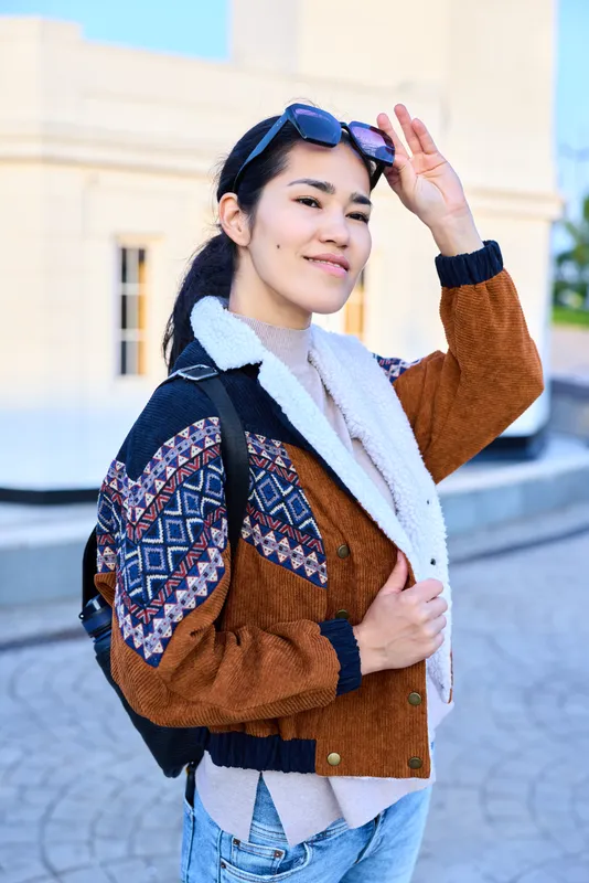 Casual street style featuring a patterned brown jacket, backpack, sunglasses, and a confident pose in an outdoor urban setting, conveying fashion and travel vibe