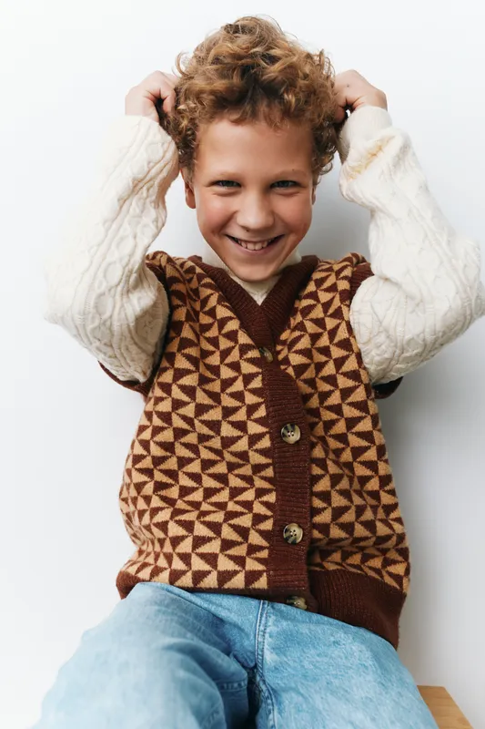 Young boy wearing a geometric brown knit vest over a cream sweater, grinning while adjusting his curly hair, set against a clean neutral backdrop for a bright, friendly look.