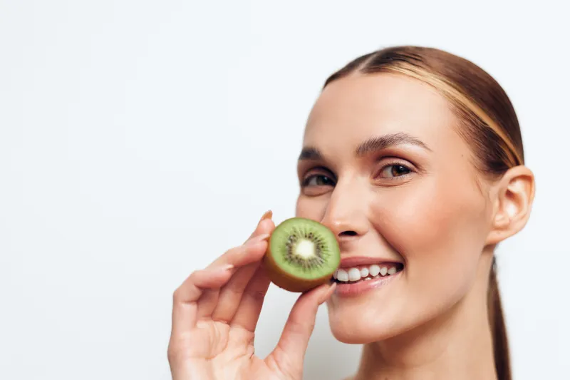 Smiling woman holding a fresh kiwi slice near her face, natural light, clear skin, healthy fruit, white background, close up, joyful expression, clean beauty concept