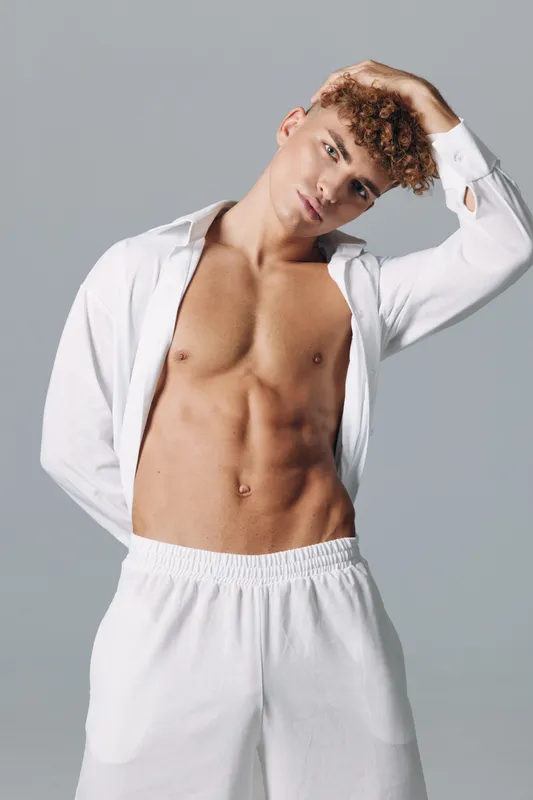 Young man with curly hair and bare torso posing in white open shirt and shorts cleaning sweat underarm featuring athletic body on neutral studio background close up portrait.