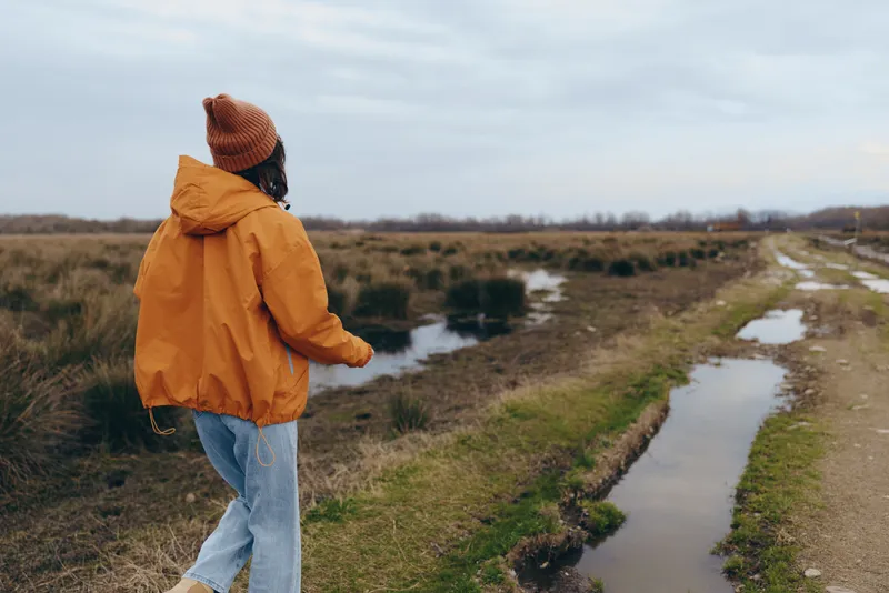 woman lifestyle walking outdoors along river trail, countryside fields and grass, orange jacket and knit hat, back view on wet path, cloudy sky, peaceful nature travel, relaxation