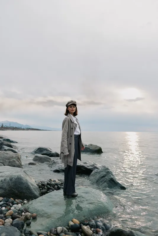 Young woman in casual outfit stands on rocky shore near calm sea under cloudy sky. Concept of solitude, nature, and peaceful outdoor lifestyle with soft natural light.