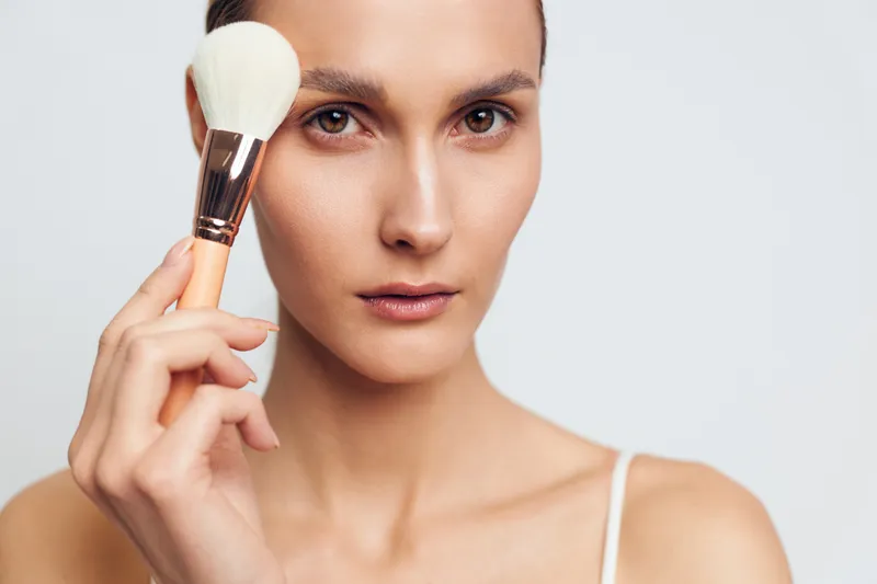 A young woman with clear skin holds a makeup brush near her face against a plain background. The image represents skincare, beauty routine, and natural makeup application.