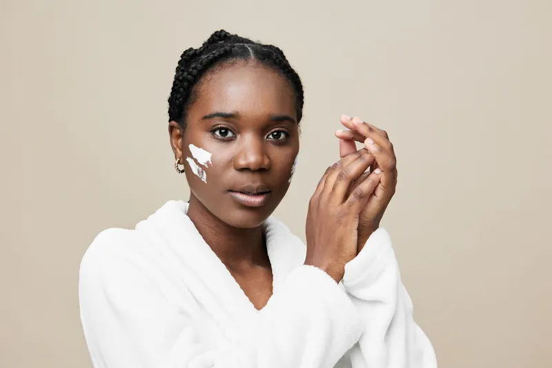 Young Black woman applying skincare cream in a white robe with a neutral background representing self care and beauty routines