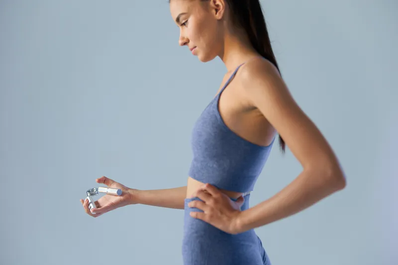 Woman sport fitness in blue activewear using a hand gripper for strength training, focused and determined, isolated on a soft grey background with minimalist style.