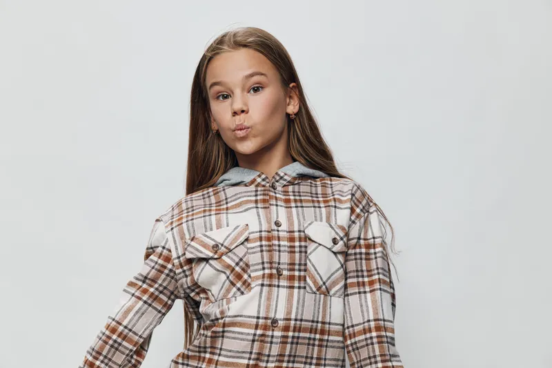 Girl with long hair in checkered shirt posing with playful expression on white background. Casual young female showing fun and confident attitude in studio photo session for lifestyle.