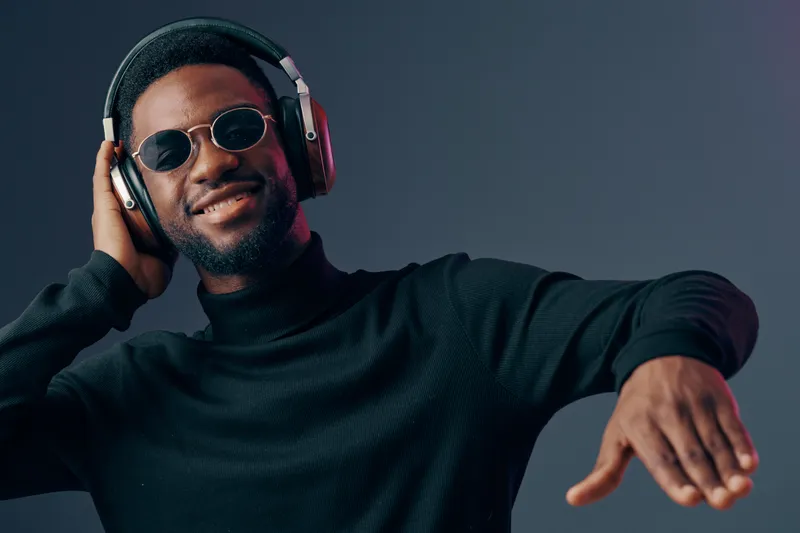 Happy African man enjoying music with headphones in stylish black outfit and sunglasses, gray background emphasizing a modern, cool vibe