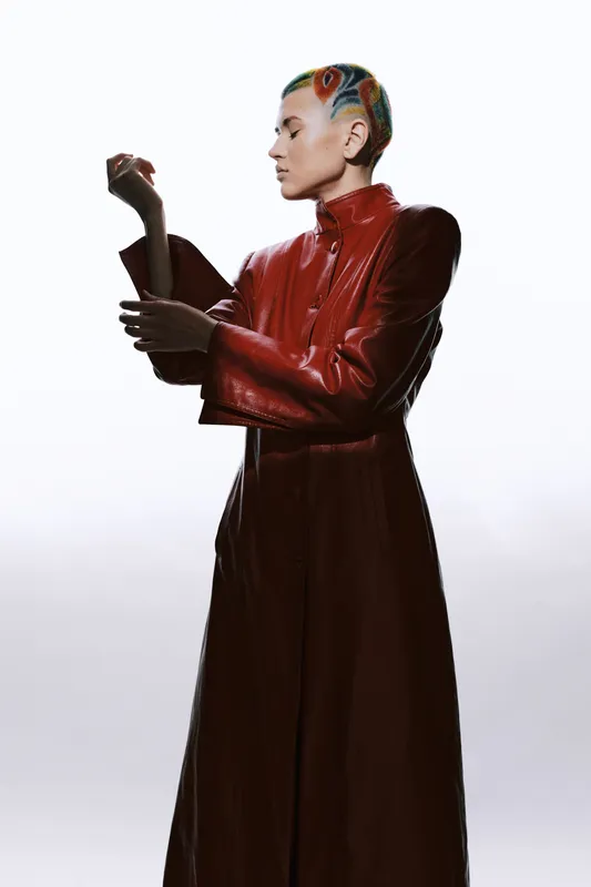 Woman with colorful braided hair in stylish red leather coat, standing in profile against a bright white background, expressing calm and modern fashion attitude.