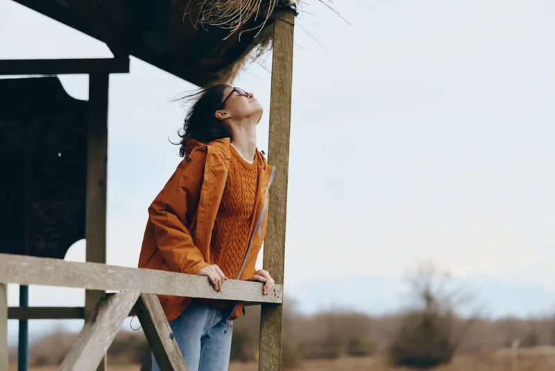 Woman lifestyle photo of a young girl in orange jacket standing by a wooden fence outdoors, she leans on the railing and looks up, calm spring air, blurred trees background, natural light