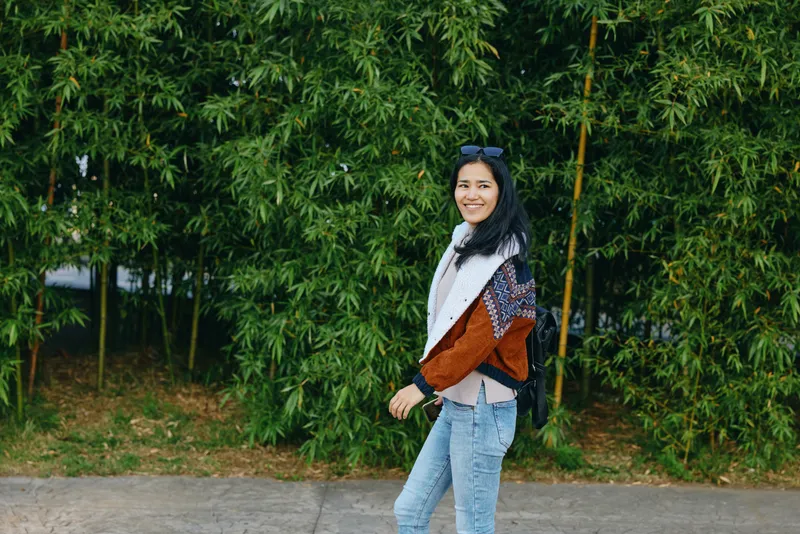 Woman in casual jacket and jeans walking outdoors along a green bamboo backdrop, sunglasses on head, smile, candid street style, fresh daytime mood and relaxed fashion vibe
